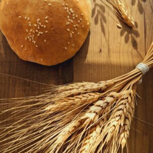 A round loaf of sesame-seed bread with wheat stalks on a wooden table, lit by warm sunlight.