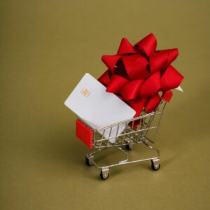 A miniature shopping cart holds a gift card adorned with a red bow, set against a green background.