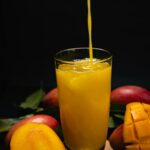 Vibrant fresh mango juice pouring into glass surrounded by ripe mangoes and leaves.